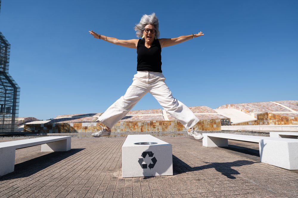 Woman with gray hair and glasses jumping energetically outdoors in an urban public space with modern architecture and clear blue sky, businesswomen, Spain.