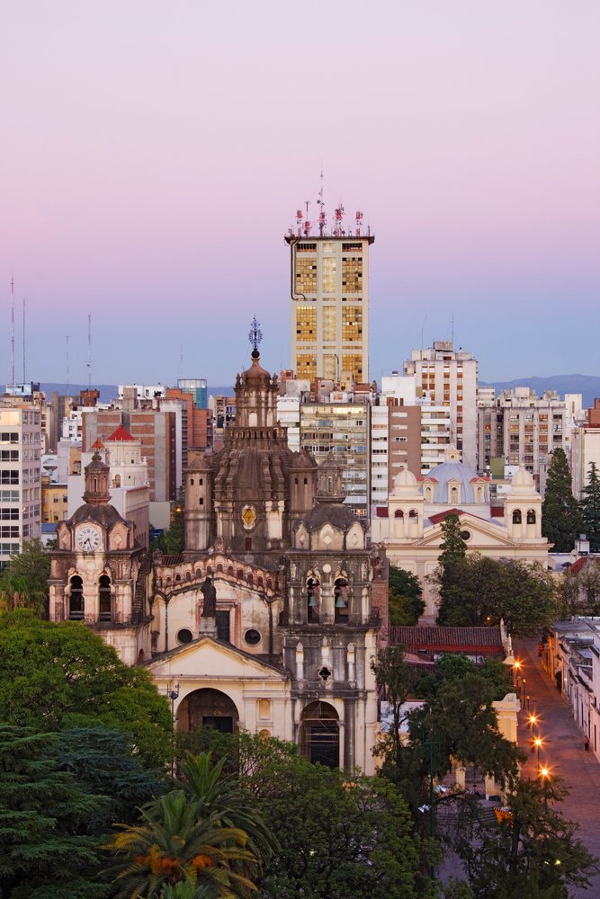 Cordoba Cathedral prominently rises within, Argentina's urban skyline at dusk with surrounding city buildings and trees.