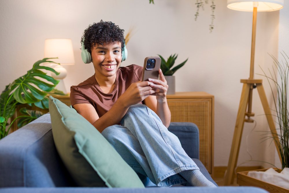 Young woman wearing headphones and holding a phone sits smiling on a blue couch in a living room with a floor lamp and monstera plant.