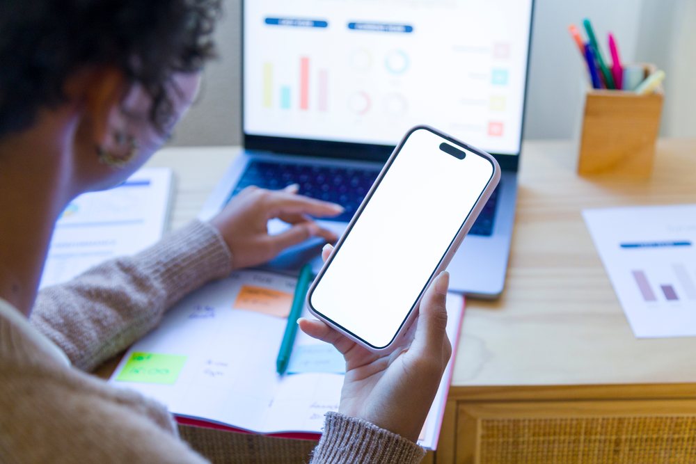 Female working remotely at home office desk using smartphone and laptop with visible business charts, remote work.