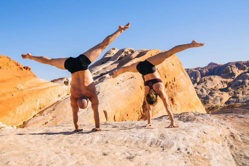 Barefoot couple performs handstands on rocky terrain beneath a clear blue sky, yoga, acrobatics, USA.