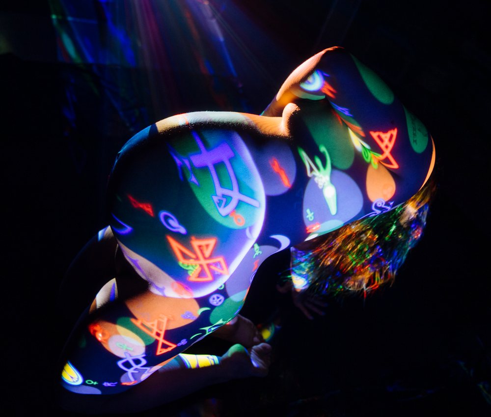Pregnant woman kneeling with colorful projection body painting on her torso against a dark background, pregnancy.