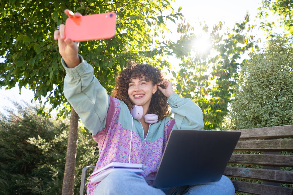 Young woman with curly hair takes a selfie on a bench outdoors with a laptop, notebook, and headphones in a sunny park, Spain.