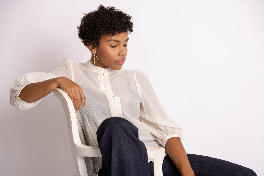 Woman with curly hair sitting relaxed on a white wooden chair wearing a white blouse, dark jeans, and gold earrings in a studio, fashion, young culture.