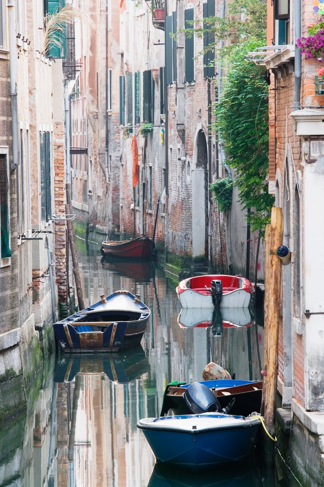 Venice canal with boats moored along a narrow waterway between brick buildings with window shutters and greenery, Italy.