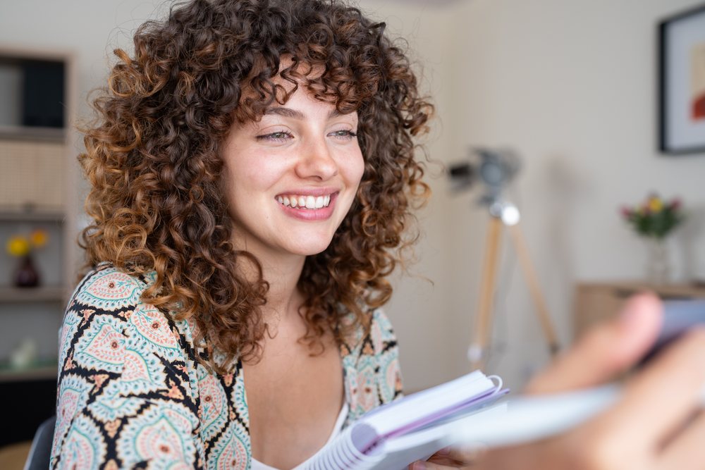 Smiling woman with curly hair holding a notebook inside a home interior setting, Spain.