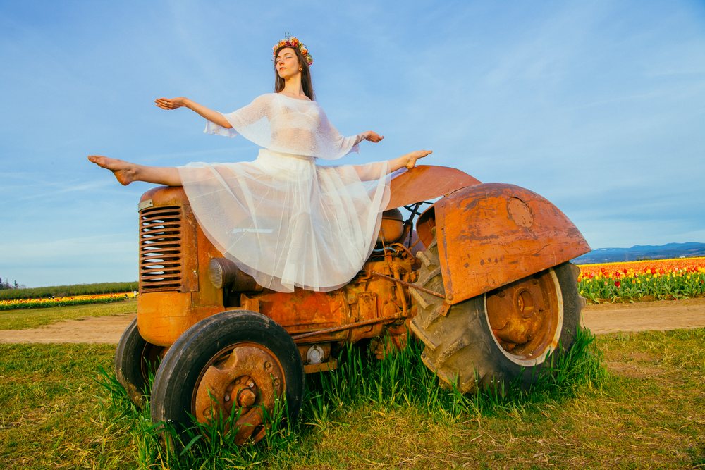 Woman wearing white dress and flower crown performs a ballet split on an old rusty tractor in a flower field, USA.