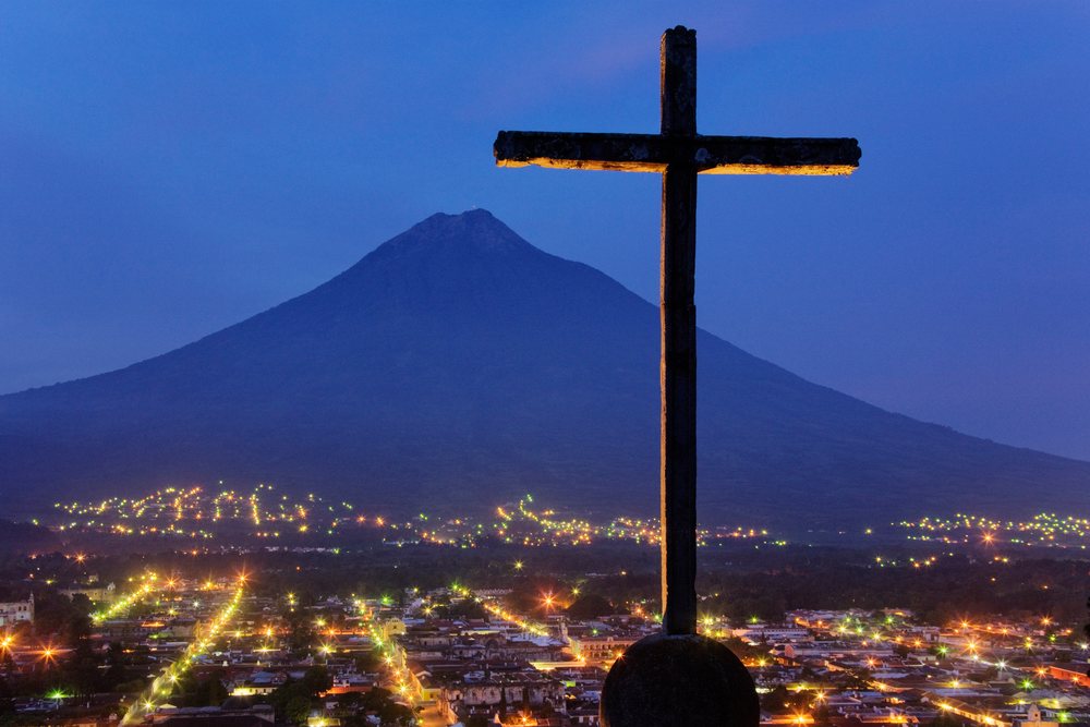 Silhouette of a cross stands before Volcan Agua mountain and city lights at dusk, Sacatepequez Department, Guatemala.