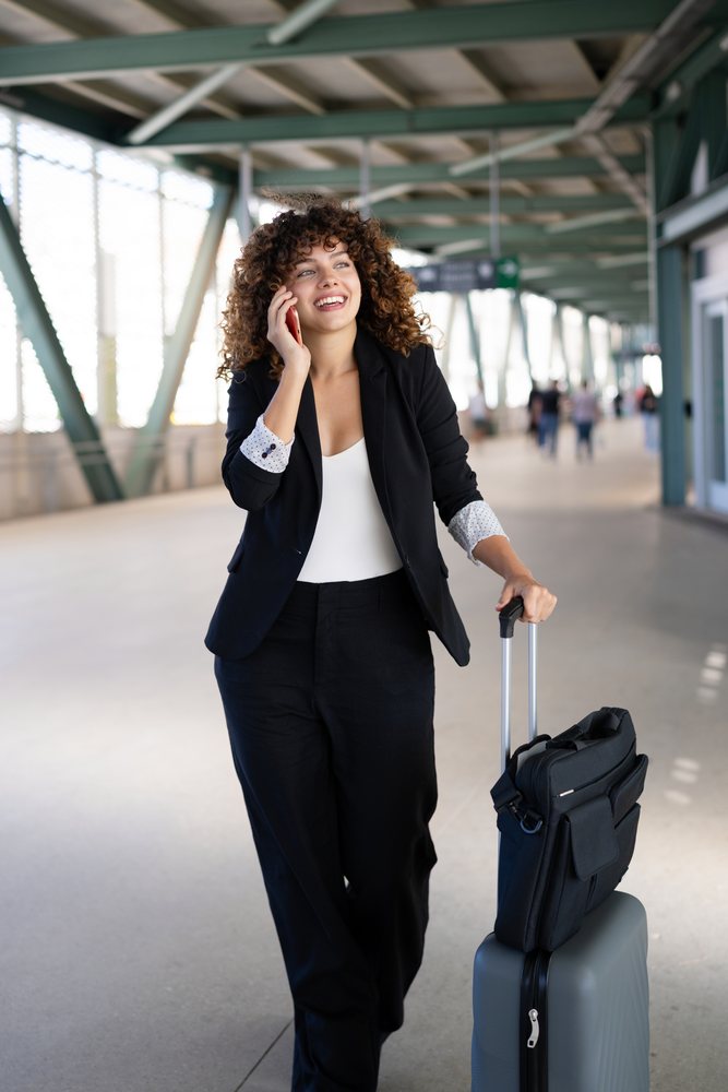 Woman with curly hair wearing business attire talks on her phone while pulling a grey suitcase with a black bag through an indoor airport corridor, Spain.
