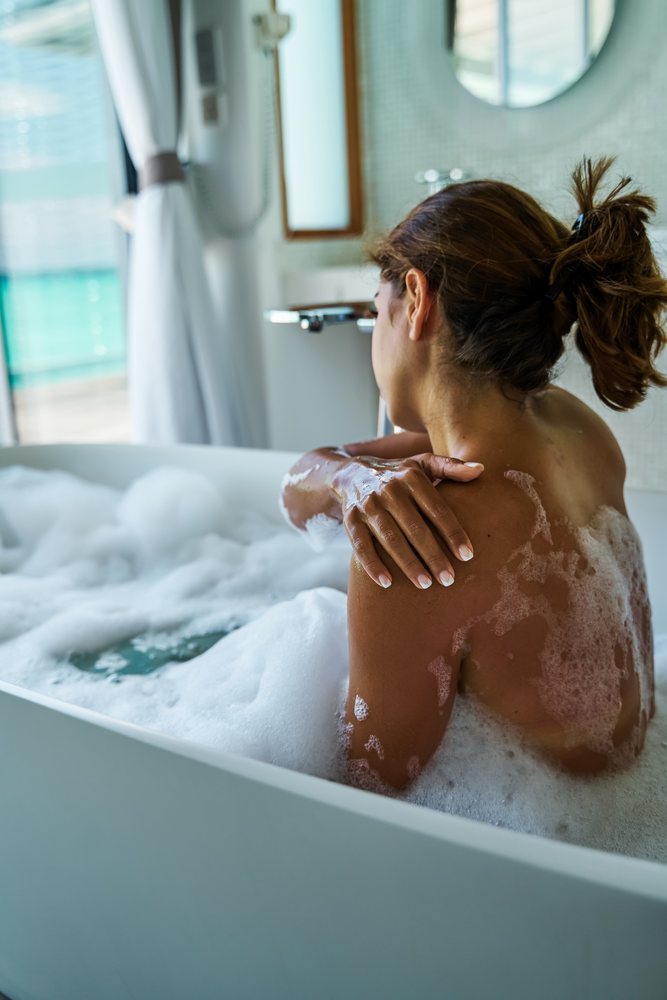 A woman is enjoying a relaxing foamy bath in a modern bathroom with white bathtub and curtains in the, Maldives.