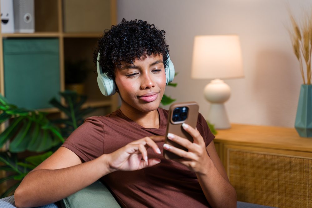 Woman with curly hair wearing headphones and holding a smartphone while sitting on a sofa in a living room with houseplants and a lamp, Spain.