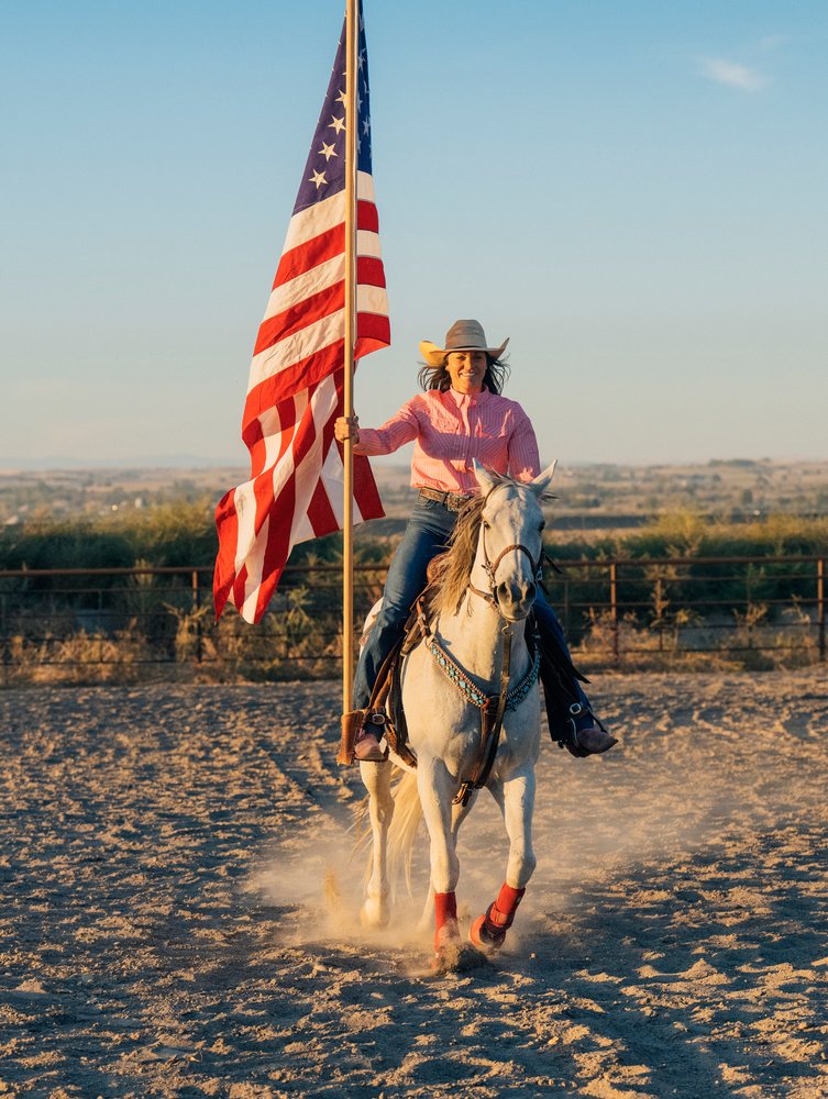 Woman wearing a cowgirl hat rides a white horse holding an American flag outdoors at sunset, Southwest USA.