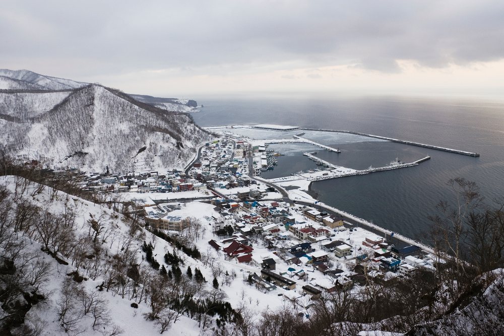 Rausu town and harbor on the snow-covered Shiretoko Peninsula coast with mountains under a cloudy winter sky, Japan, Asia.