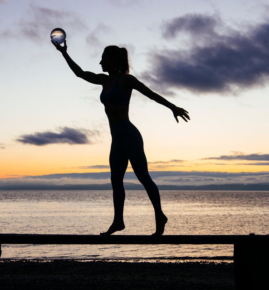 Silhouette of a woman balancing on a beam holding a crystal ball on the beach at sunset, WA, USA.