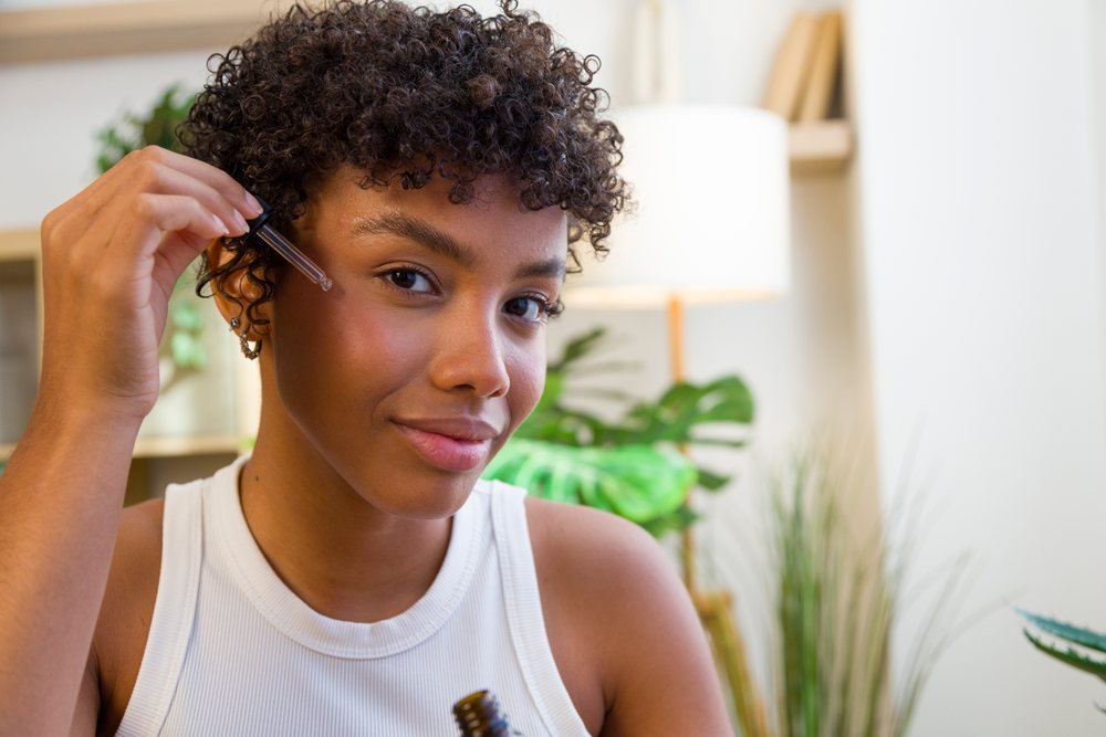 Young woman applying serum to her face during a skincare routine at home with plants and a floor lamp in the background.