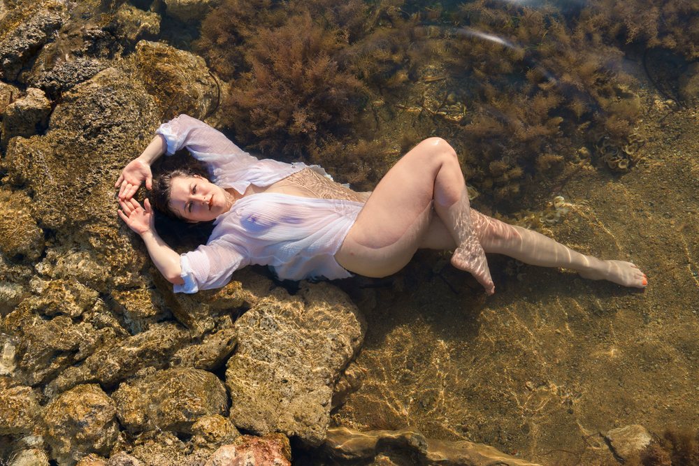 Woman lying in shallow water among rocks and seaweed, relaxing in sunlight wearing a wet white shirt, photo shoot, Ibiza, Spain.
