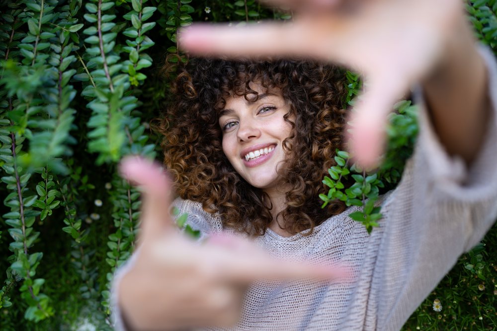 Woman with curly hair smiling and framing her face with hands outdoors in greenery, home story, influencer, Spain.