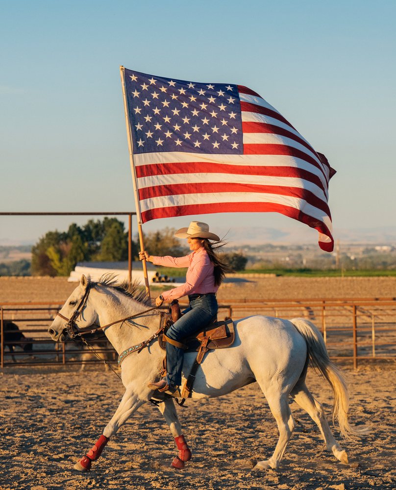 Woman riding a white horse waving an American flag in an outdoor arena at sunset, Southwest USA.