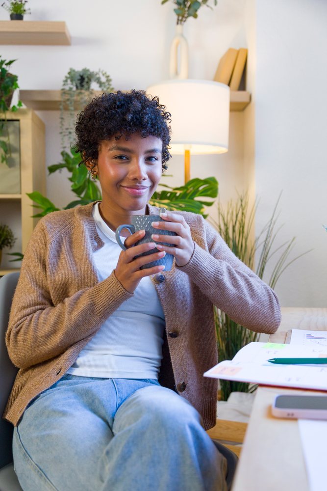 Woman sitting in a home office holding a mug and smiling with plants and books in the background.