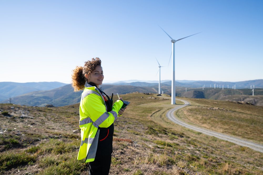 Woman engineer wearing yellow safety jacket holding notepad and pen at a wind farm amid rolling hills under clear blue sky, sustainability, renewable energy, Galicia, Spain.