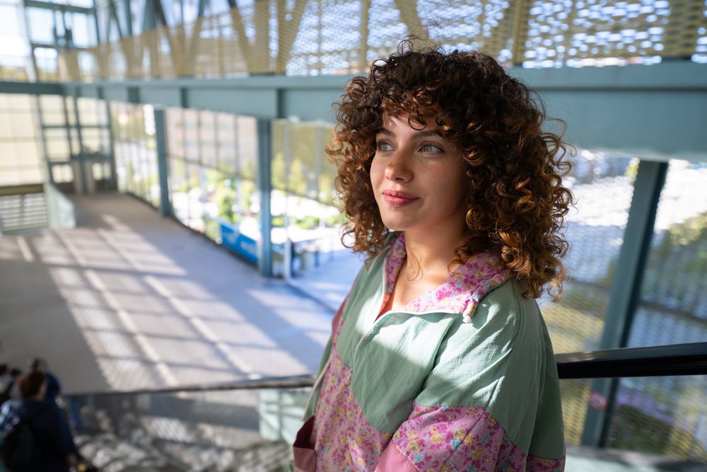 Woman with curly hair wearing a floral light jacket stands thoughtfully on an outdoor staircase in sunlight in an urban setting, Spain.