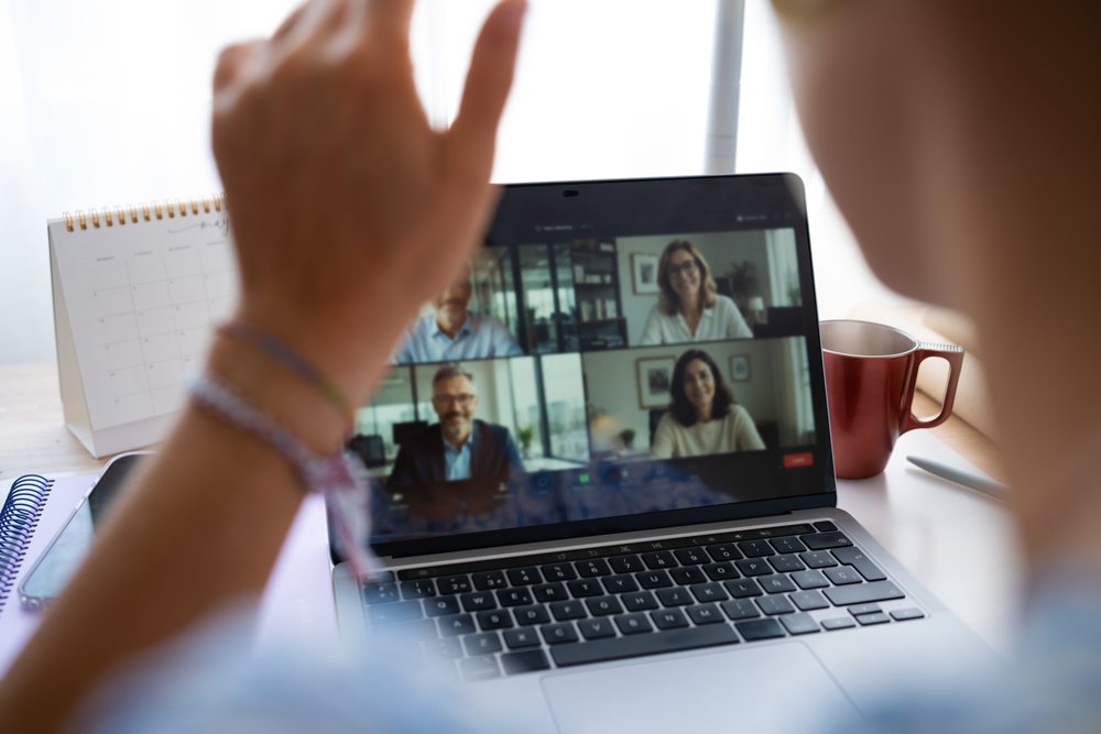 Person waving during a video conference on a laptop in a home office with a calendar and coffee mug nearby, telecommuting, Spain.