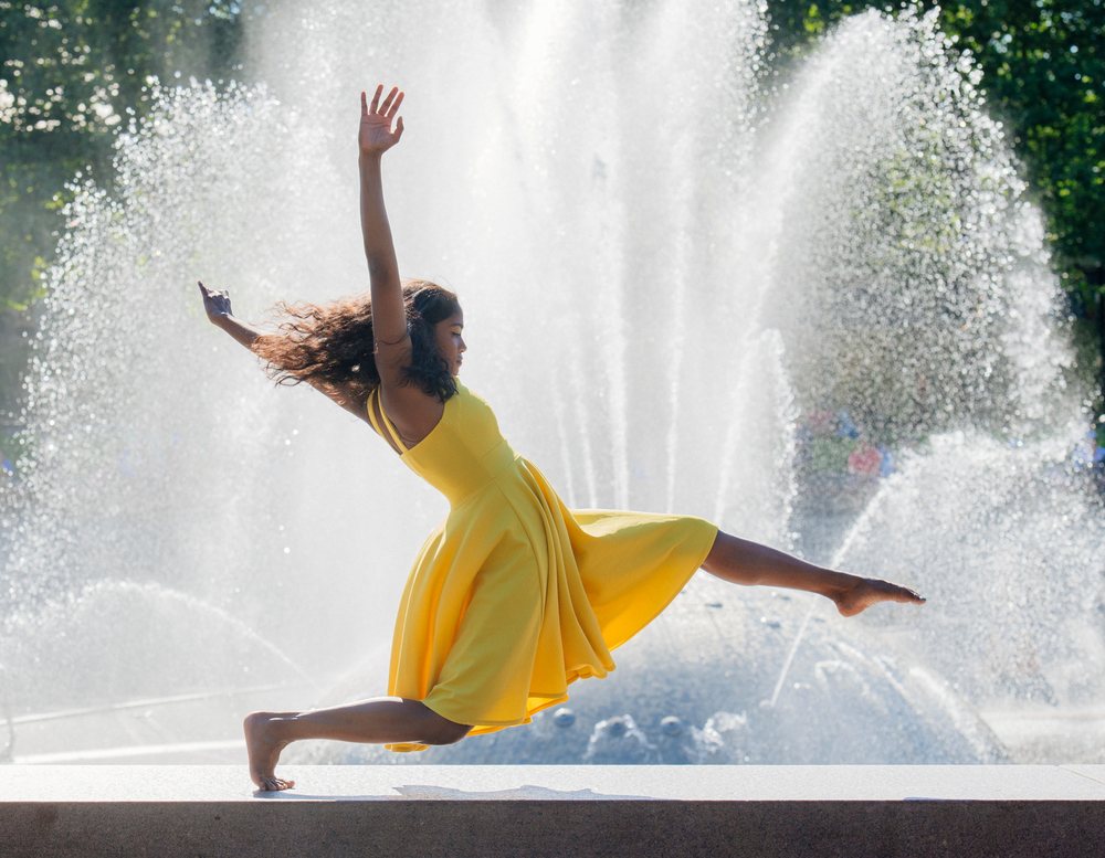 Woman in a yellow dress leaps barefoot gracefully beside a large outdoor fountain spraying water in sunlight, USA.