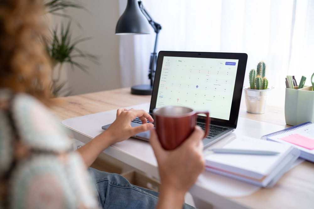 Woman holding a coffee mug and using a laptop displaying a calendar at her home office desk, Spain.