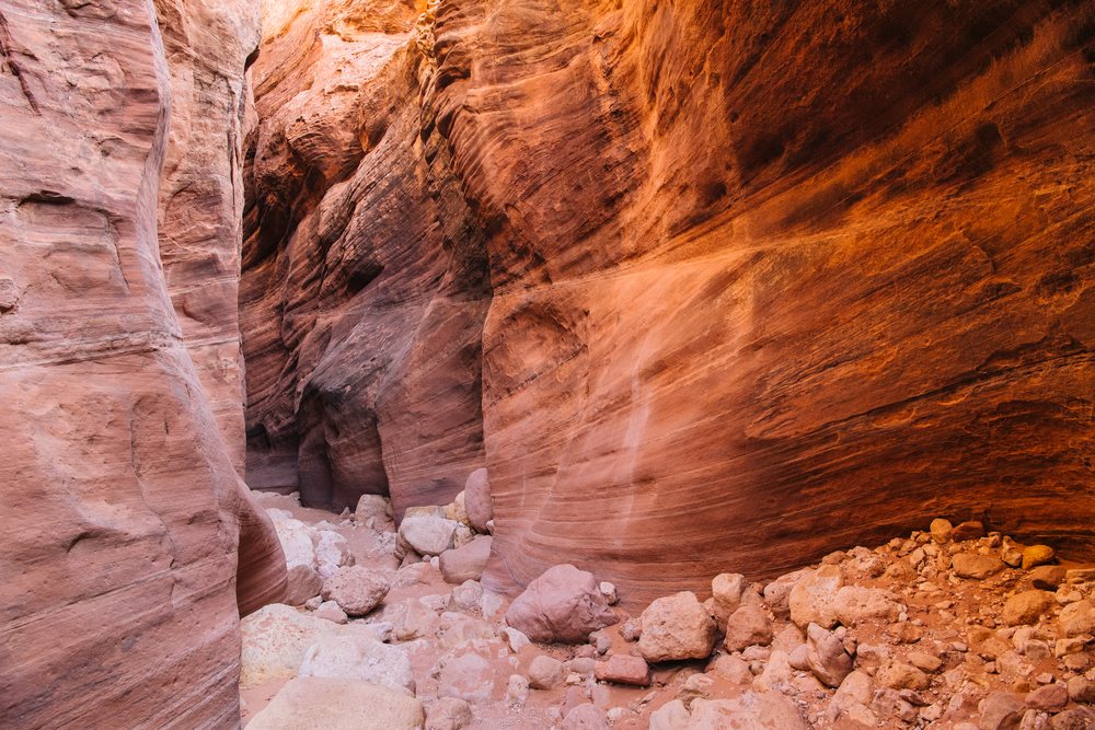 Slot canyon in Utah desert featuring layered red sandstone rock walls and scattered sandstone boulders under natural sunlight, Buckskin Gulch, USA.