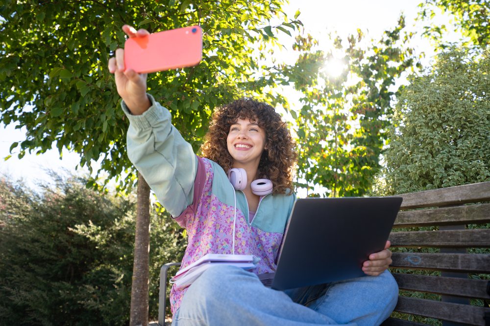 Woman with curly hair smiles while taking a selfie outdoors on a bench holding a laptop and notebooks in a sunny park, Spain.