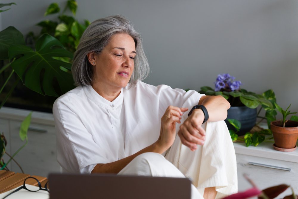 Middle-aged woman wearing white clothes checking her wrist device indoors near houseplants, Spain.