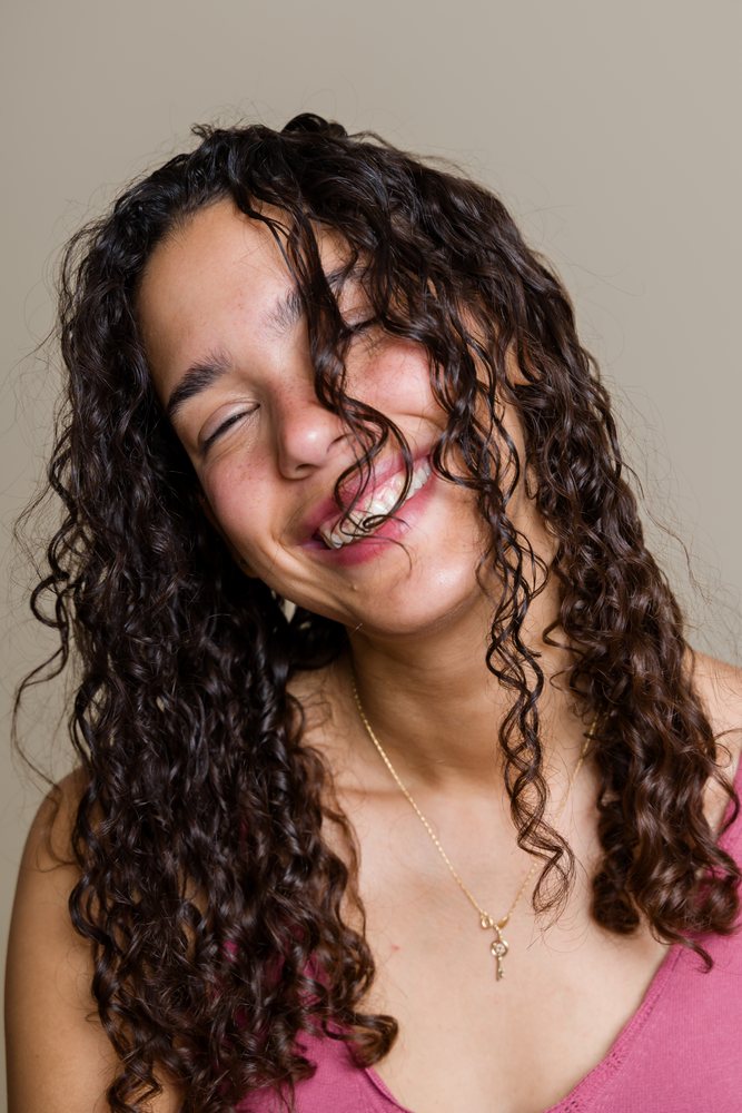 Young adult woman with long curly brown hair smiles joyfully with closed eyes wearing a necklace and pink top in a studio portrait taken, beauty shoot, Spain.