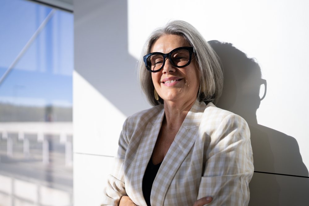 Woman with gray hair and black glasses smiles indoors with crossed arms in natural light, businesswoman, traveler, Spain, Madrid.