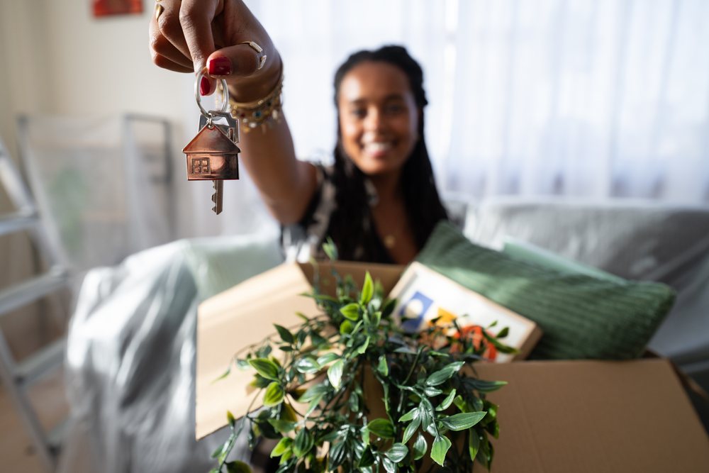 A woman holding a house key with a house-shaped keychain while unpacking boxes in a living room during moving day, Spain.