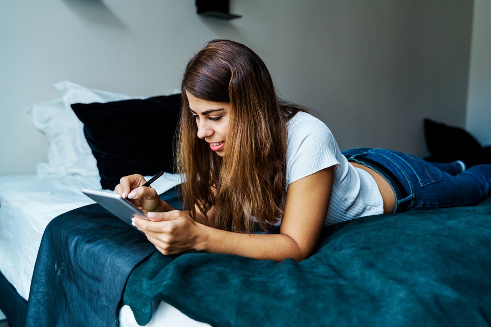 A woman uses a tablet and stylus on a bed