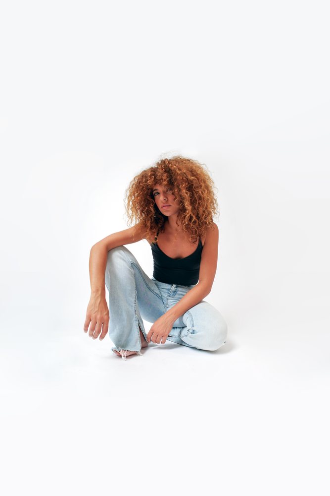 Woman with curly hair sitting barefoot on the floor wearing a black tank top and light blue jeans in a studio, Malaga, Spain.