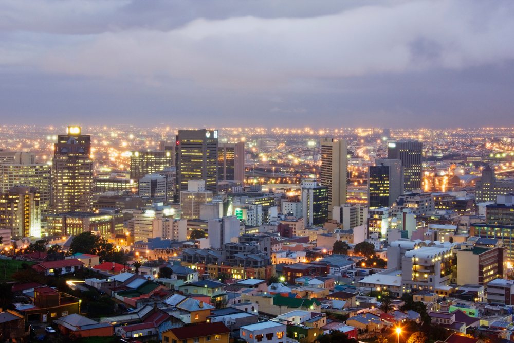 Dusk cityscape of Cape Town showing illuminated buildings, residential houses, and road lighting under a cloudy evening sky, Western Cape, South Africa.