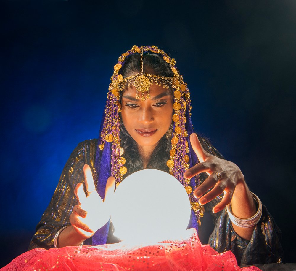 A woman dressed as a fortune teller gazes intently into a glowing crystal ball in a dark indoor setting in the, USA.