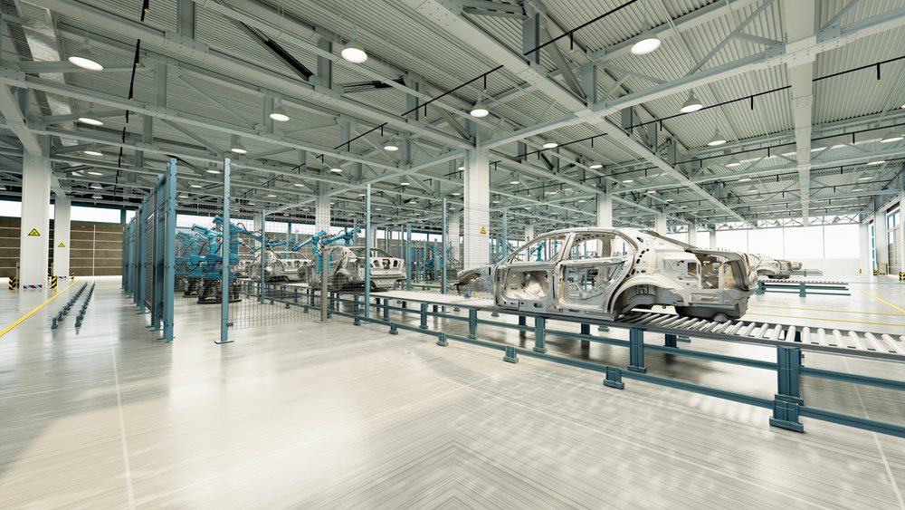 Car frames move along conveyor belts in a large, automated automotive manufacturing factory interior with robotic arms and assembly stations.
