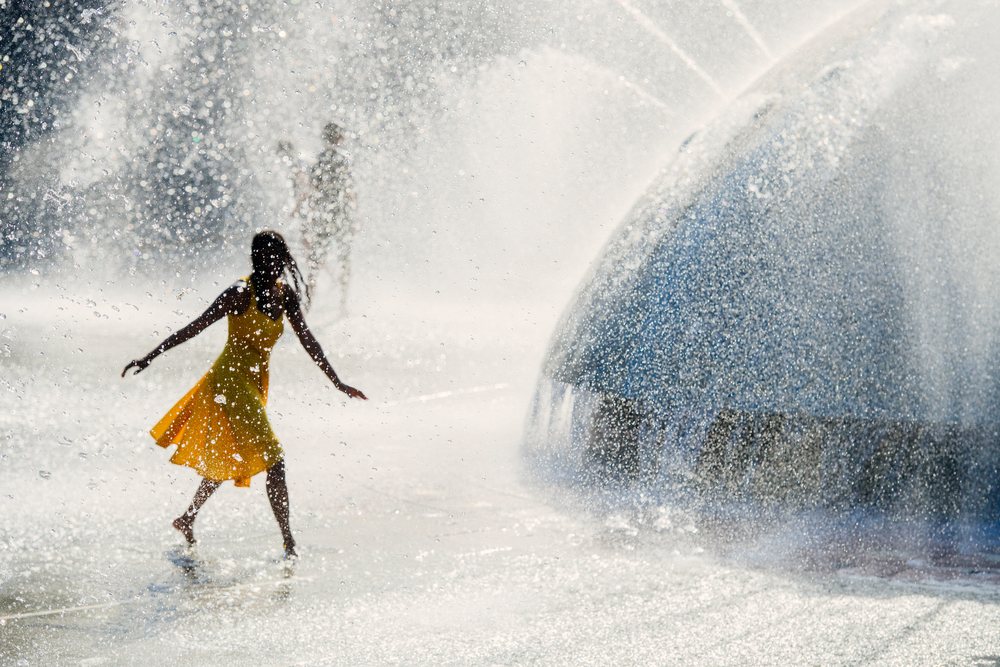 Woman in a yellow dress walks barefoot through spraying water at a public park fountain in the USA on a sunny summer day.