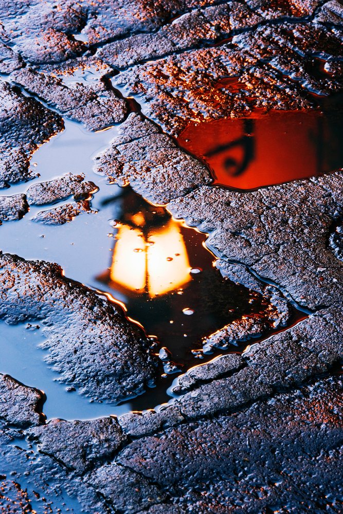 Glowing street lamp reflects in a puddle on wet, textured asphalt during evening.