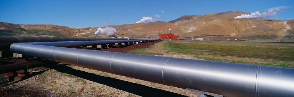 Large metal pipeline extends through a volcanic landscape with steam vents visible under a clear blue sky.