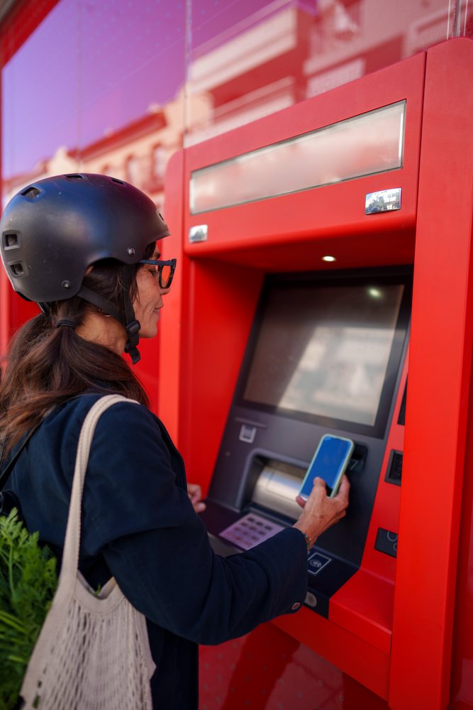 Woman wearing a black helmet uses a smartphone at a red ATM outdoors in Málaga during daylight.