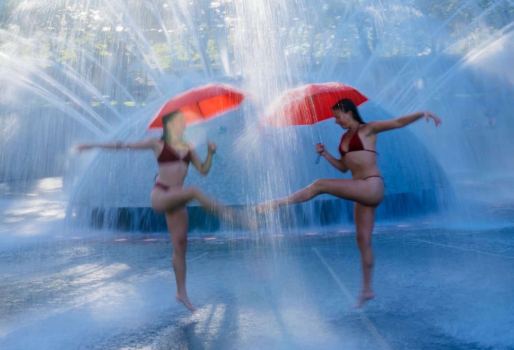 Two women dancing and balancing with red umbrellas in a water fountain, engaging in playful acrobatics and yoga, USA.