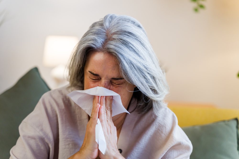 Woman with gray hair sneezing into a tissue while seated on a couch in an indoor living room setting, Spain.