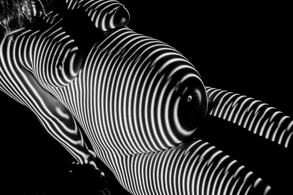 Pregnant woman with striped black and white light pattern on her nude torso against a black background, pregnancy.