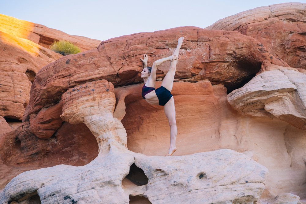 Woman performs yoga acrobatics while stretching barefoot on red rock formations outdoors, USA.