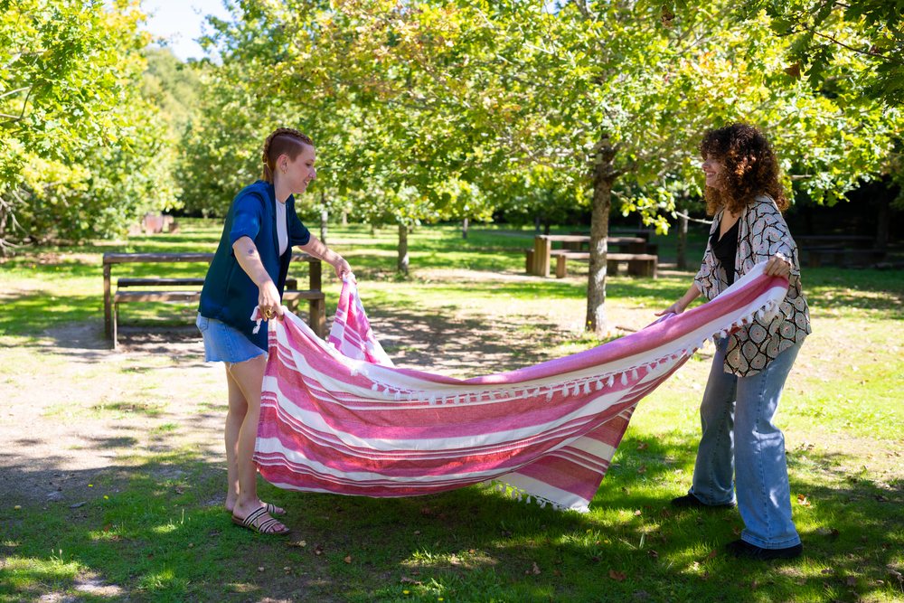 Two women spreading a striped picnic blanket in a sunny green outdoor park with trees and benches, Spain.