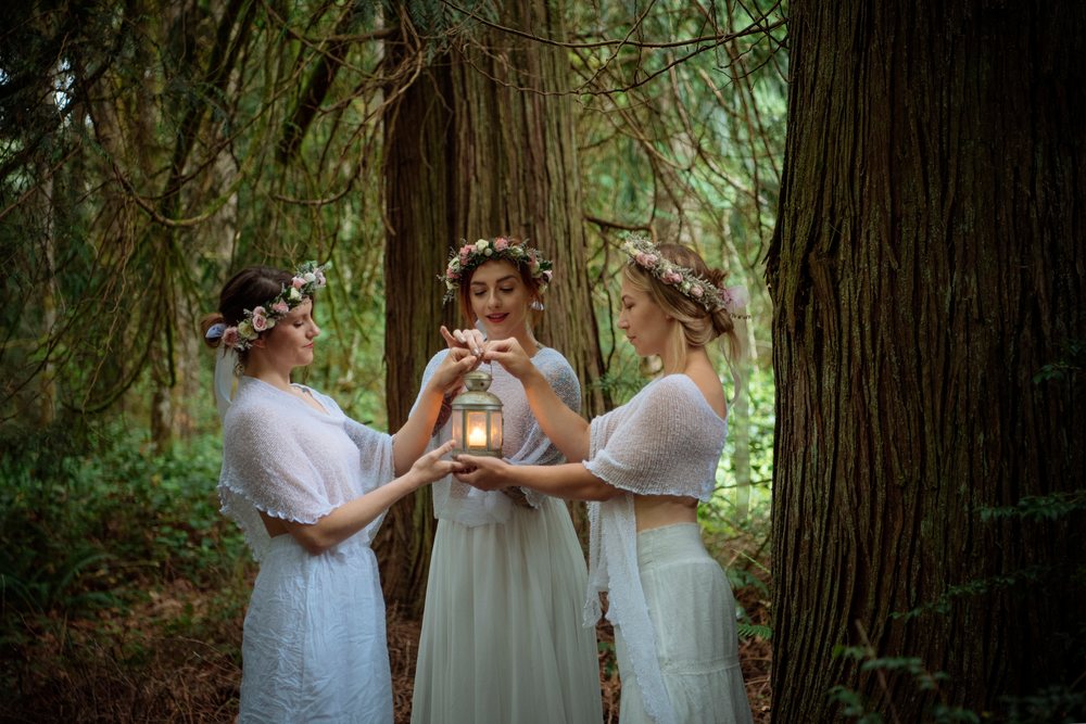 Three women wearing flower crowns hold a lit lantern together in a peaceful forest, muses, USA.