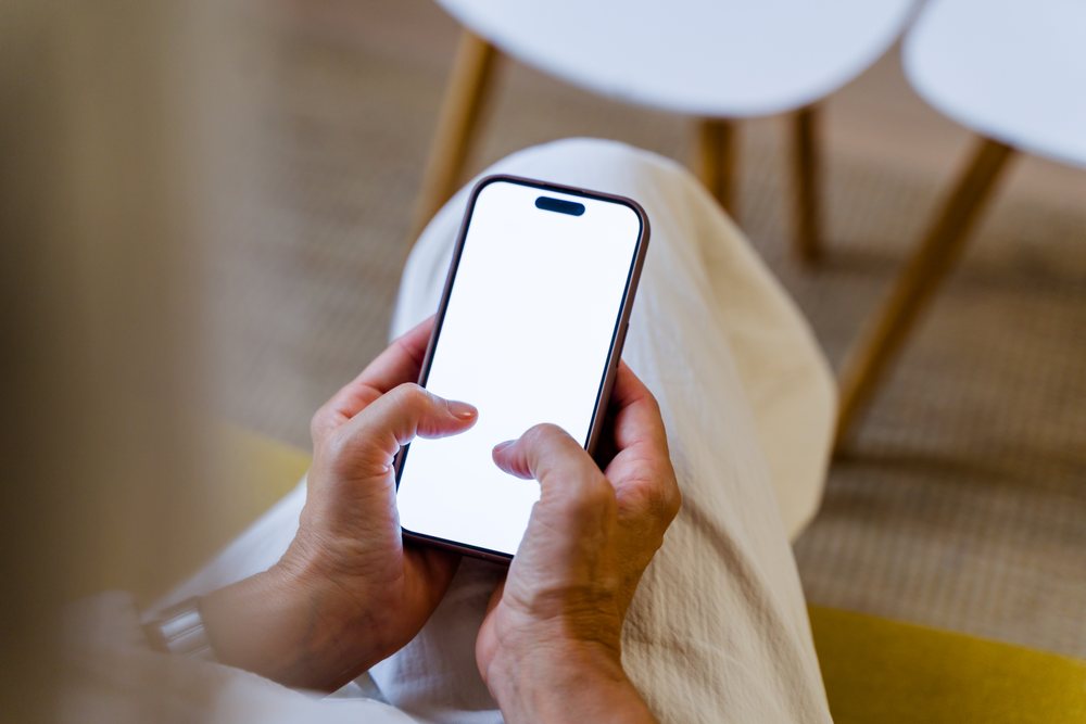 Person sitting and holding a smartphone with a white blank screen in a modern setting, Spain.